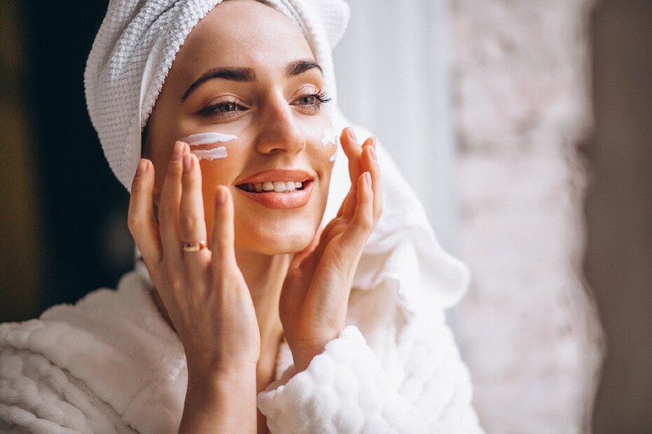Mujer de frente aplicándose crema hidratante en el rostro, sobre productos de belleza que no pueden faltar en tu rutina diaria