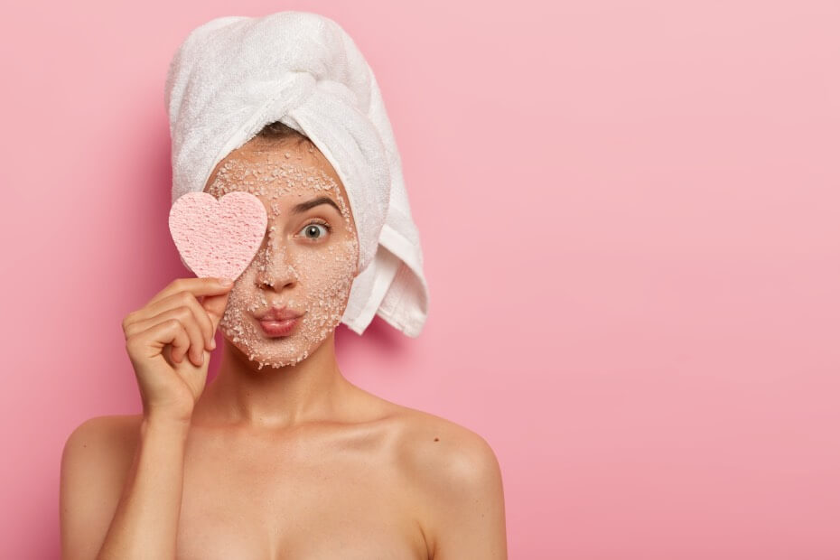 Mujer recién bañada con turbante en el pelo aplicándoselos una mascarilla facial sosteniendo un corazón de papel sobre fondo roza